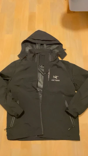 Svart vindjacka från Arc'teryx - Svart vindjacka från Arc'teryx med huva och dragkedja. Jackan har vita detaljer och logga på bröstet, samt reflekterande prickar på axlar och rygg. Tillverkad i slitstark polyester som är vatten- och vindtät. Perfekt för utomhusaktiviteter och äventyr.