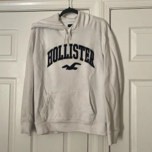 Vit hoodie från Hollister med tryck - Vit hoodie från Hollister med stort mörkblått broderat logotryck på bröstet och klassisk känguruficka. Tröjan har huva med snörning och långa ärmar. Perfekt för chill dagar och enkel att matcha med jeans eller mjukis.