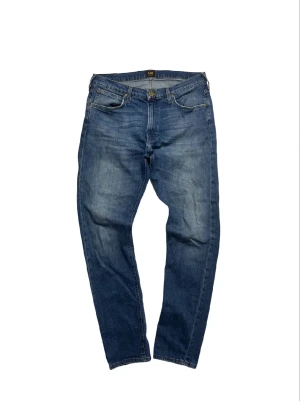 Blå Lee Luke jeans W34 L32 - Snygga blå jeans från Lee i modellen Luke, med klassisk femficksdesign och brunt läderpatch bak. Jeansen har en rak passform och är tillverkade i slitstarkt bomullsjeansmaterial. Perfekt för dig som gillar en tidlös och clean look.