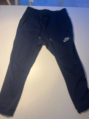 Mörkblå mjukisbyxor från Nike - Snygga mörkblå mjukisbyxor från Nike med klassisk logga på benet. Byxorna har snörning i midjan, två sidofickor med dragkedja och ribbade muddar vid bensluten. Perfekta för chill eller träning, riktigt sköna och stilrena.