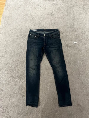 Mörkblå Jack n Jones jeans - Snygga mörkblå Jack n Jones  jeans med klassisk femficksdesign och smal passform. Jeansen har diskreta slitningar framtill och silverfärgade knappar. Det är Slim/Glenn som passform i storleken 32/32! Bara höra av er vid ev frågor eller funderingar!