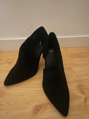 Svarta mocka pumps från H&M - Snygga svarta pumps från H&M i storlek 40, tillverkade i mjuk mocka. Skorna har en spetsig tå och låg klack som ger en elegant och modern look. Perfekta för dig som gillar stilrena och trendiga skor.