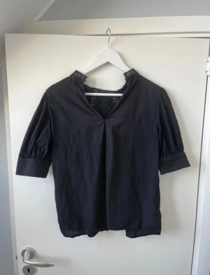 Svart blus med spetsdetaljer - Säljer en svart blus med v-ringning och halvkort puffärm. Blusen har fina spetsdetaljer vid halsen och över axlarna, samt dekorativa knappar vid ärmsluten. Materialet känns lätt och luftigt, perfekt för dig som gillar en stilren look med lite extra detaljer.