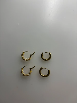 Två par guldfärgade örhängen - Säljer två par snygga guldfärgade örhängen. De med pärlor är färgat silver. Ett par är klassiska hoops i guldton, det andra paret har en unik design med små vita pärlor längs hela ringen. Perfekt för dig som gillar att mixa och matcha smycken för en personlig stil.