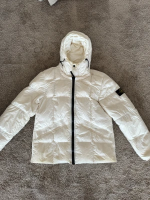 Vit dunjacka från Stone Island - Säljer en fet vit dunjacka från Stone Island med huva och svart dragkedja. Jackan har quiltad design, två snedställda fickor och klassisk Stone Island patch på ärmen. Perfekt för kalla dagar och har en clean look som sticker ut.