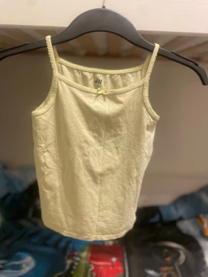 Ljusgul linnetopp från H&M med rosett - Söt ljusgul linnetopp från H&M i storlek 122/128. Toppen har smala axelband, dekorativ spetskant och en liten rosett framtill. Materialet är mjuk bomull och toppen är armlös, perfekt för varma dagar. Barn till tjejer 