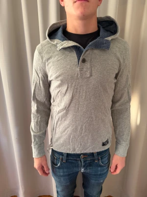 Grå hoodie Abercrombie & Fitch XS - Snygg grå hoodie från Abercrombie & Fitch i storlek XS. Tröjan har en mjuk huva med blått foder, halv knappslå framtill och långärmad passform. Perfekt för chill dagar och enkel att matcha med jeans. Materialet är bomull och polyester för skön känsla.