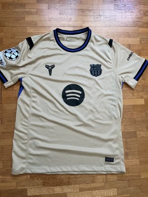 Beige FC Barcelona Rashford tröja - Säljer en beige FC Barcelona fotbollströja från Nike med svarta och blå detaljer. Tryck med Spotify-logga, FCB-emblem och Rashford 14 på ryggen. Champions League-märke på ärmen och UNHCR-tryck nedtill. Materialet är lätt och andas, perfekt för match eller träning.