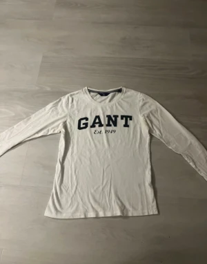 Vit långärmad tröja från GANT - Vit långärmad tröja från GANT med stor mörkblå logotyp och texten 'Est. 1949' på bröstet. Rund halsringning med ribbad kant och mjukt bomullsmaterial. Perfekt för dig som gillar klassisk och enkel stil med tydlig branding.