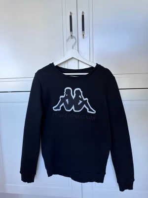 Svart sweatshirt Gosha Rubchinskiy x Kappa - Svart sweatshirt med Kappa-logga i vitt och ryska bokstäver broderade under. Tröjan har rund hals, långa ärmar och ribbade muddar. Perfekt för dig som gillar streetwear och vill sticka ut med en unik collab.
