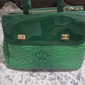 Grön handväska från Tom&Eva Paris - Snygg grön handväska med lyxig känsla från Tom&Eva Paris. Väskan har glansig ovandel, guldfärgade metalldetaljer och strukturmönstrad framsida som påminner om struts. Praktiskt innerfack med dragkedja och stadiga handtag.