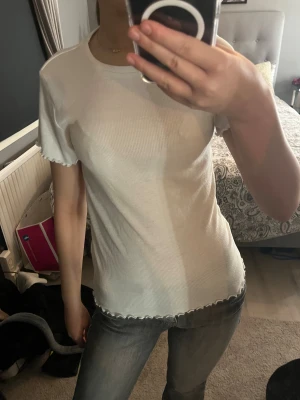 Vit ribbad topp från 157 stl XS - Säljer en vit ribbad topp från 157 i storlek XS. T-shirten har korta ärmar och söta volangdetaljer vid ärmslut och nederkant. Materialet är mjukt och stretchigt, perfekt för en chill och enkel look. Rund halsringning och slim passform.