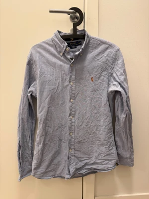 Blå skjorta Polo Ralph Lauren Slim Fit - Snygg ljusblå skjorta från Polo Ralph Lauren i slim fit-modell. Klassisk button-down krage, broderad logga på bröstet och vita knappar. Tillverkad i bomull och har långa ärmar. Perfekt för en clean och stilren look.