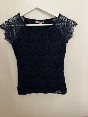 Mörkblå spetsblus från KappAhl XS - Säljer en mörkblå blus från KappAhl i storlek XS. Blusen har korta ärmar och är helt täckt av vågigt mönster. Den är figurnära och har rund halsringning. Perfekt för sommaren då den är lätt och tunn.