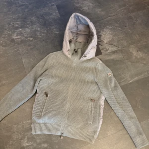 Moncler Cardigan - Snygg grå pufferjacka från Moncler med stickade ärmar och huva. Jackan har dragkedja framtill, två fickor med dragkedja och ribbade muddar. Baksidan och huvan är quiltad och fylld med dun, medan ärmarna är stickade för extra komfort. Perfekt för kyliga dagar.