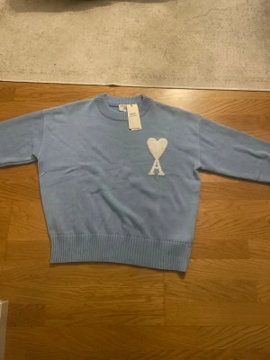 Ljusblå stickad tröja från Ami - Supersnygg ljusblå stickad tröja från Ami med vit hjärtlogga och A på bröstet. Tröjan har rund hals, ribbade muddar och är långärmad. Tillverkad i mjuk bomull, perfekt för dig som gillar stilrena detaljer och vill ha något som sticker ut.