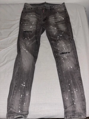 Grå Dsquared2 jeans med färgstänk - Snygga grå jeans från Dsquared2 med coola vita färgstänk och slitna detaljer på benen. Modellen har klassisk femficksdesign, raka ben och låg midja. Jeansen är tillverkade i bomull och har Dsquared2-logga på etiketten och vid gylfen.