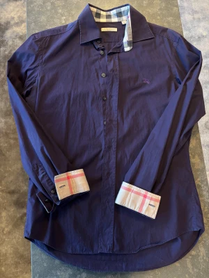 Mörklila skjorta från Burberry Brit - Snygg mörklila skjorta från Burberry Brit med klassiskt rutigt mönster på insidan av krage och manschetter. Skjortan har långa ärmar, knappar framtill och broderad logga på bröstet. Tillverkad i bomull för en skön känsla och stilren look.