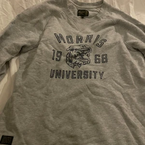 Grå sweatshirt Morris University - Grå sweatshirt från Morris med tryck i blått framtill, där det står 'Morris University 1968' och en bulldog-logga. Tröjan har långärmad design och rund hals. Perfekt för dig som gillar college-stil och vill ha en chill look.