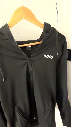 Svart kofta från Hugo Boss - Svart vindjacka från Hugo Boss med stilren design och logga på bröstet. Jackan har huva, hel dragkedja framtill och praktiska fickor. Materialet är lätt och syntetiskt, perfekt för dig som gillar sportig och clean look. Snygga detaljer med BOSS-tryck på dragkedjan.