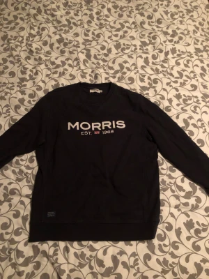 Svart sweatshirt från Morris, M - Svart sweatshirt från Morris i storlek M med vit broderad logga och texten 'MORRIS EST. 1968' på bröstet. Tröjan har rund hals, ribbade muddar och långa ärmar. Materialet är mjuk bomull som känns skön mot huden. Fler bilder kan lösas bara att höra av sig och pris kan diskuteras vid snabb affär.