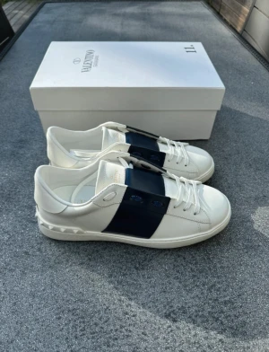Valentino Garavani Open sneakers vit/blå - Snygga Valentino Garavani Open sneakers i vitt skinn med bred marinblå läderdetalj över mitten. Klassisk rund tå, vita snören och ikoniska nitar på hälen. Perfekta för dig som gillar exklusiva och stilrena sneakers med en twist.