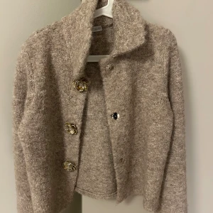 Beige cardigan jacka med blomknappar - Säljer en beige cardigan jacka från Zara Knit med fluffig, stickad känsla och unika blommiga knappar i guldton. Jackan har lång ärm, bred krage och croppad passform som ger en cool och trendig look. Perfekt för dig som gillar detaljer och vill sticka ut. Ej använd.