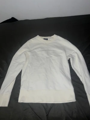 Vit sweatshirt från Woolrich - Vit sweatshirt från Woolrich med broderad logga framtill. Tröjan har rund hals, ribbade muddar och är långärmad. Tillverkad i mjuk bomull som är skön mot huden. Perfekt för dig som gillar clean och enkel stil.