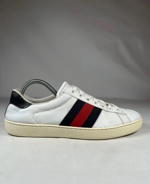 Gucci vita sneakers med röd/blå rand - Snygga vita sneakers från Gucci med klassiska ränder i rött och mörkblått på sidan. Skorna har rund tå, snörning och en beige platt sula. Ovandelen är i skinn och hälen har svart detalj. Perfekt för dig som gillar stilrena och lyxiga sneakers. Skriv om ni har frågor eller funderingar!😊