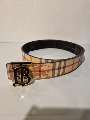 Rutig Burberry bälte med guldspänne - Snyggt bälte från Burberry med klassiskt rutigt mönster i beige, brunt och rött. Bältet har ett stort, guldigt spänne med Burberrys ikoniska B-logga. Perfekt accessoar för att lyfta din outfit med en lyxig touch.