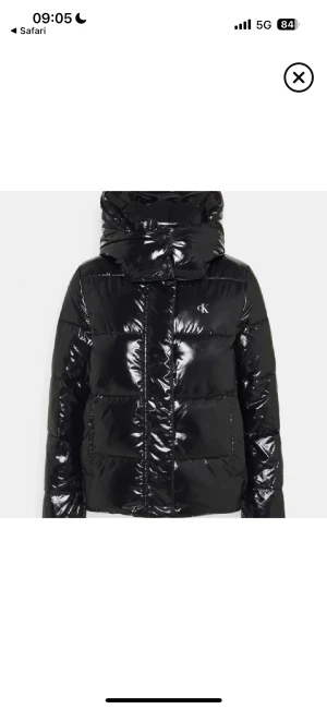 Svart pufferjacka från Calvin Klein - Snygg svart pufferjacka från Calvin Klein med glansig finish och hög krage. Jackan har en diskret CK-logga på bröstet, praktiska fickor och dragkedja framtill. Perfekt för kalla dagar när du vill ha både stil och värme.
