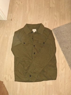 Olivgrön overshirt från Selected homme - Säljer en olivgrön overshirt från Selected Homme med klassisk krage, två bröstfickor med knappar och raka ärmar. Jackan är i bomull och har en clean, avslappnad look som passar perfekt till jeans eller chinos. Hör av er vid frågor eller prisförslag🫱🏼‍🫲🏽