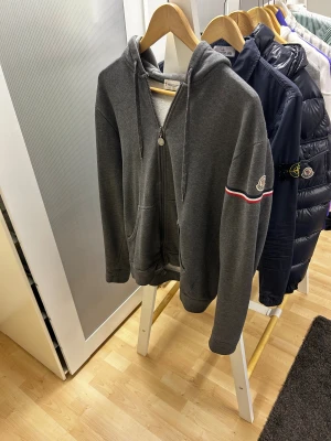 Moncler Maglia Cardigan - Snygg grå hoodie från Moncler med huva och hel dragkedja framtill. Tröjan har långa ärmar, två fickor och en broderad Moncler-logga på vänster ärm med röd, vit och blå rand. Perfekt för chill dagar och streetwear vibes. Storlek L fits M.