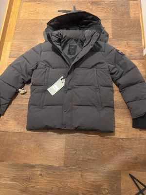  Canada Goose jacks grå - Säljer en mörkgrå dunjacka från Canada Goose med huva och stora fickor framtill för ett bra pris. Jackan har quiltad design, ribbade muddar och klassisk patch på ärmen. Perfekt för kalla dagar när du vill hålla stilen och värmen. 