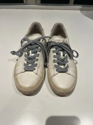 Vita Veja sneakers med grå snörning - Säljer ett par klassiska Veja sneakers i vitt skinn med grå snörning och beige detaljer på hälen. Snygg rund tå och platt sula, perfekt för dig som gillar stilrena och minimalistiska sneakers. Veja-loggan syns på sidan och hälen.