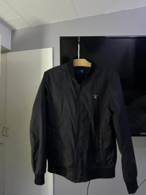 Svart bomberjacka från GANT, S - Svart bomberjacka från GANT i storlek S. Jackan har ribbade muddar, dragkedja framtill och klassisk rund krage. Insidan har tryck med 'New Haven 49 GANT Connecticut'. Materialet är glansigt och känns lätt, perfekt för dig som gillar stilren och sportig look.