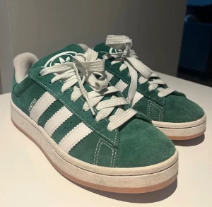 Adidas Campus gröna sneakers mocka - Adidas Campus sneakers i grön mocka med vita detaljer och klassiska tre ränder på sidan. Skorna har vit platt sula, rund tå och snörning. Adidas logga på plösen och hälen. Fräscha och stilrena sneakers som lyfter din streetwear-look.