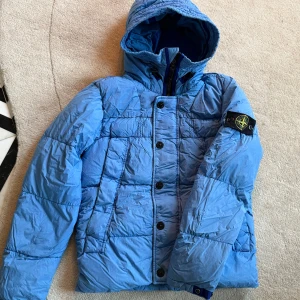 Blå dunjacka från Stone Island - Säljer en blå dunjacka från Stone Island med huva, quiltad design och ikonisk patch på ärmen. Jackan har både dragkedja och knappar framtill, samt praktiska fickor. Perfekt för dig som gillar streetwear och vill ha en snygg jacka med coola detaljer.
