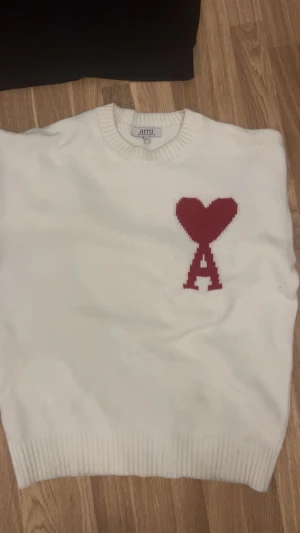 Vit stickad tröja från Ami med hjärta - Säljer en vit stickad tröja från Ami med röd hjärtdetalj och bokstaven A på bröstet. Tröjan har rund hals och ribbade muddar vid ärmslut och nederkant. Perfekt för dig som gillar stilrena plagg med en twist.