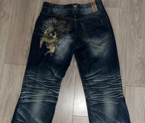 BIGTRAIN JEANS  - Säljer ett par mörkblå jeans med coolt guld- och svart drakbroderi på bakfickan. Jeansen har markerade slitningar och tvättade detaljer över benen för en edgy look. Passformen är loose och materialet är klassisk jeans. Perfekt för dig som vill sticka ut. 