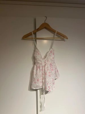 Vit blommig linnetopp med snörning - Supersöt vit linnetopp med rosa blommigt mönster och broderade detaljer. Toppen har smala axelband, v-ringning och snörning framtill. Materialet är lätt och luftigt, perfekt för varma dagar. Den har en omlottliknande passform och är ärmlös för en fräsch look.