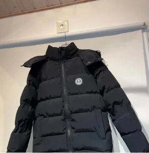 Svart pufferjacka med huva - Svart pufferjacka med huva och hög krage, perfekt för kalla dagar. Jackan har en rund logotyp på bröstet, två sidofickor och dragkedja framtill. Den är quiltad och har en clean, modern look som funkar till streetwear och chill outfits.