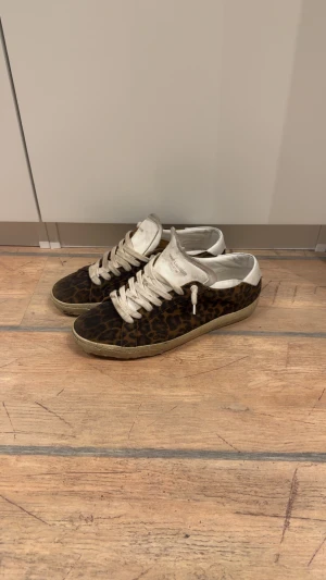 Saint Laurent leopardmönstrade sneakers - Snygga sneakers från Saint Laurent med leopardmönstrad mocka i bruna och svarta toner. Skorna har vit platt sula, rund tå och vita snören samt detaljer i vitt skinn vid häl och plös. Perfekt för dig som vill sticka ut med en cool och lyxig vibe.