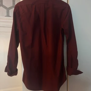 Vinröd skjorta från Polo Ralph Lauren - Säljer en vinröd skjorta från Polo Ralph Lauren i custom fit. Skjortan har klassisk krage, vita knappar framtill och lång ärm. Broderad logga på bröstet. Tillverkad i bomull och passar perfekt för dig som gillar stilrena plagg med snygg passform.