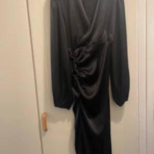 Svart omlottklänning i satin - Supersnygg svart omlottklänning i glansigt satinmaterial. Klänningen har långa puffärmar, v-ringning och snygg rynkdetalj i sidan. Perfekt för dig som vill sticka ut med en elegant och trendig look.