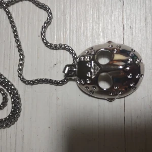 Halsband med hockey mask-hänge i silverfärg - Unikt halsband med ett coolt maskformat hänge i blank silverfärg. Hänget har detaljerade nitar och stora ögonhål, inspirerat av hockeymask eller skräckfilm. Kedjan är robust och matchar hängets metalliska look. Perfekt för dig som gillar edgy accessoarer.