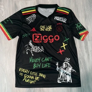 Ajax x Bob Marley Adidas fotbollströja S - Unik svart Ajax fotbollströja från Adidas med Bob Marley-tema, tryckta citat och illustrationer i vitt, rött, gult och grönt. Kortärmad med reggaefärgade ränder på ärmarna. Materialet är lätt polyester med Aeroready-teknologi. Perfekt för dig som gillar fotboll och musik.