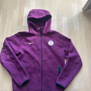 Lila PSG hoodie från Nike, stl M - Säljer en lila hoodie från Nike med Paris Saint-Germain-logga på bröstet. Tröjan har hel dragkedja, huva och två fickor med dragkedja. Perfekt för dig som gillar sportig stil och vill sticka ut med en snygg färg.