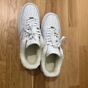 Nike Air Force 1 vit fårskinnsfodrad - Nike Air Force 1 sneakers i helvitt skinn med mjukt fårskinnsfoder på insidan. Klassisk siluett med rund tå, perforerad tåbox och snörning. Perfekta för dig som vill ha en clean look men ändå hålla värmen under kallare dagar.