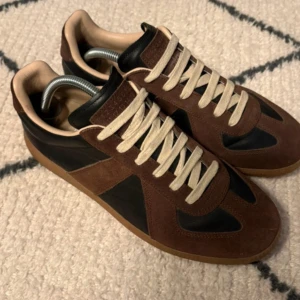 Sneakers i brun/svart - Snygga sneakers  i brunt mocka och svart skinn med beige snörning och gummisula. Klassisk siluett med rund tå och diskret logga på plösen. Perfekta för dig som gillar stilrena och trendiga skor.
